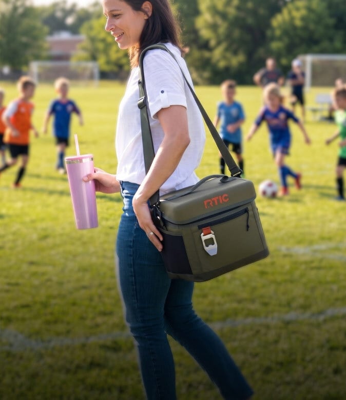 Woman carrying an olive green RTIC everyday cooler and pink tumbler while walking beside kids playing soccer on a field.