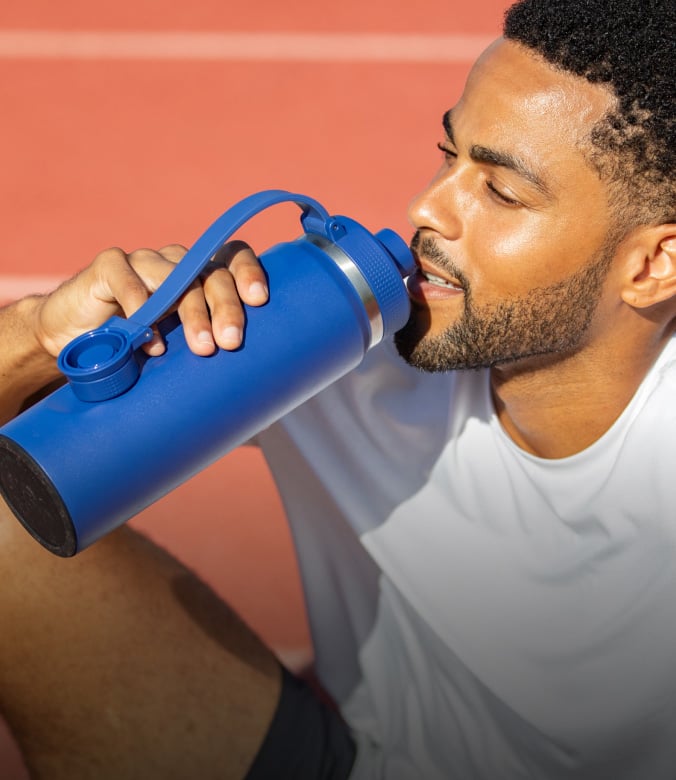 Man drinking from a large blue insulated water bottle while sitting outdoors in athletic wear under bright sunlight