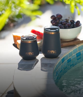 Two black soft-touch RTIC tumblers by a pool with sliced watermelon, cantaloupe, and grapes in the background on a sunny day.