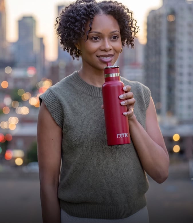 Woman smiling while holding a red RTIC water bottle against a city backdrop.
