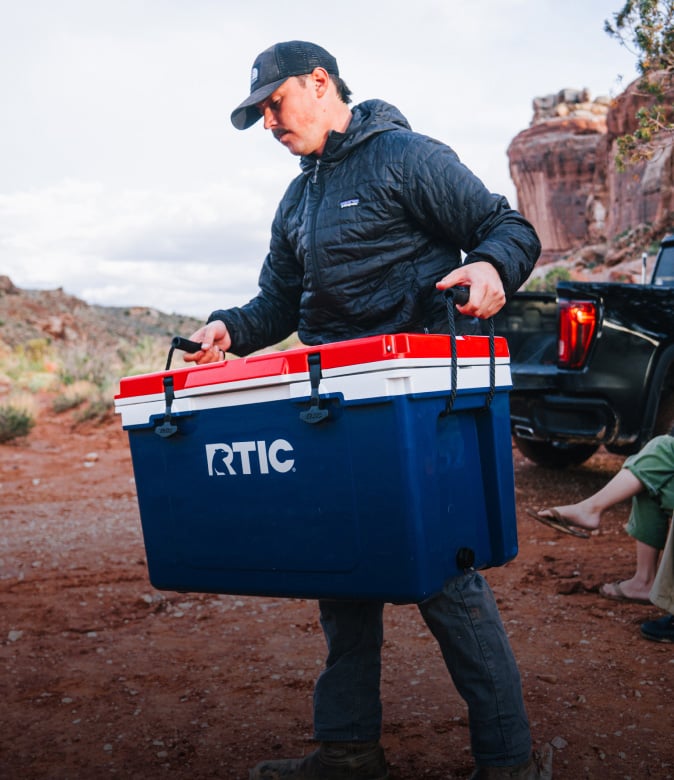 Person carrying a red, white and blue RTIC hard cooler beside a pickup truck in a rugged desert landscape.