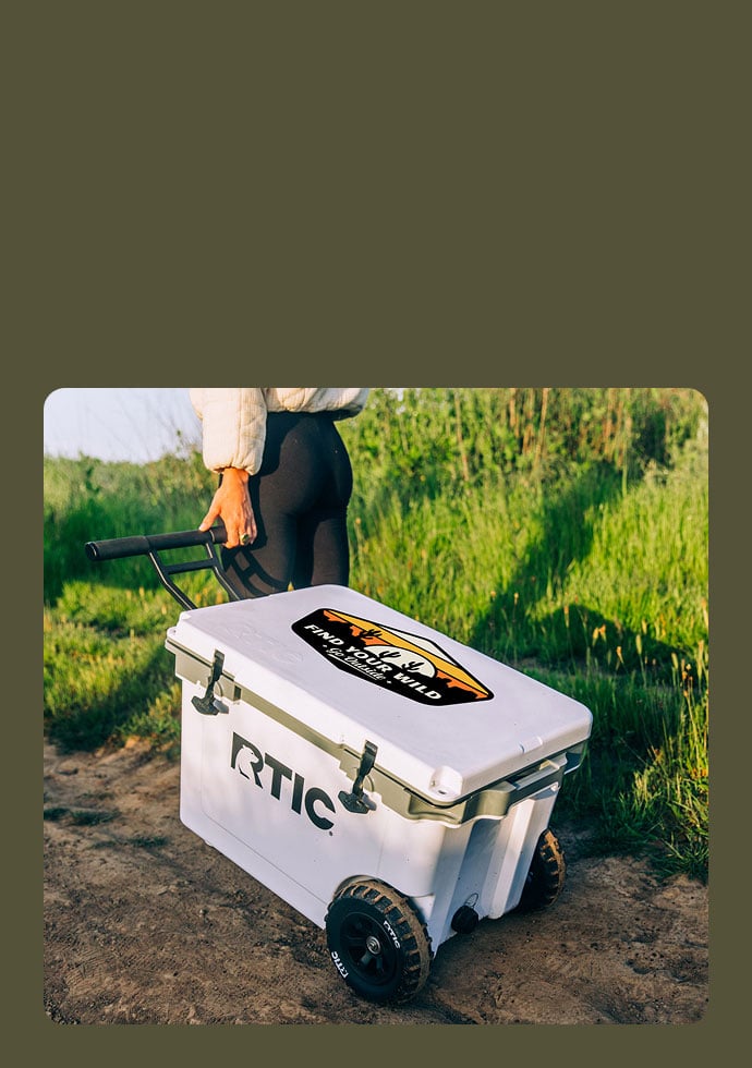 A woman pulling a personalized, white 52 QT Ultra-Light Wheeled Cooler through dirt and grass on a hill.