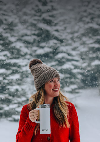 A girl smiles in snowy woods while holding an RTIC travel mug.