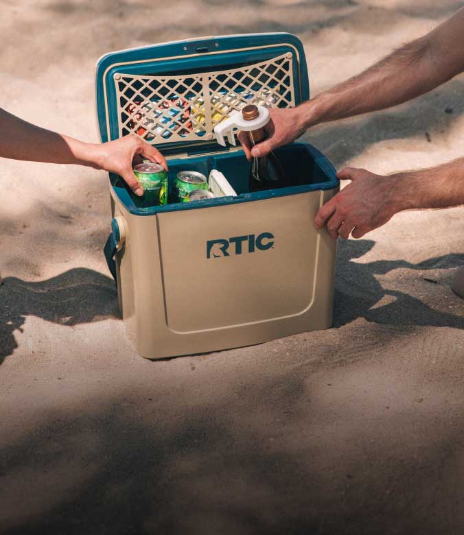 Friends sharing a tan RTIC 16 QT Road Trip Cooler on the beach.