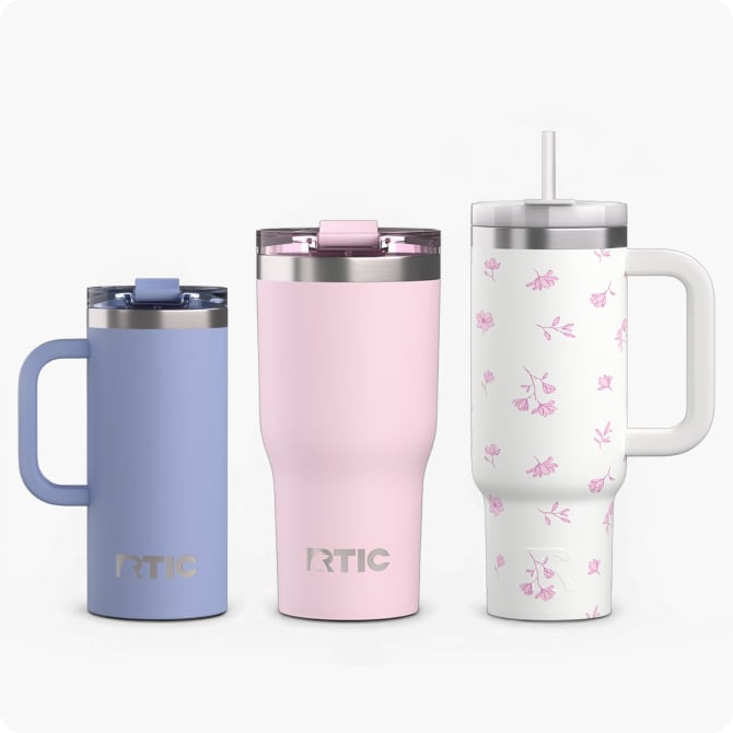 Three insulated RTIC tumblers: light purple handled mug, pink tumbler, and white floral cup with handle and straw on a neutral background.