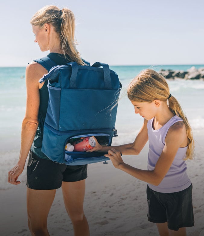 a woman carries a blue cooler backpack on a beach while a young girl zips open the lower compartment to reach inside.