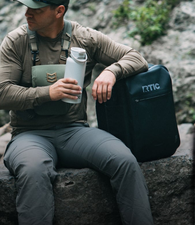 Man sitting outdoors drinking from a Bottle, leaning on a Ultra-Dry Backpack Cooler.