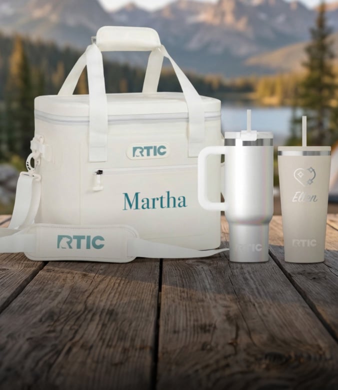 White RTIC soft cooler next to two tumblers on a wooden table by a lake and mountains.
