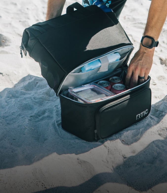 Open black RTIC cooler backpack filled with food and drinks on a sandy beach.