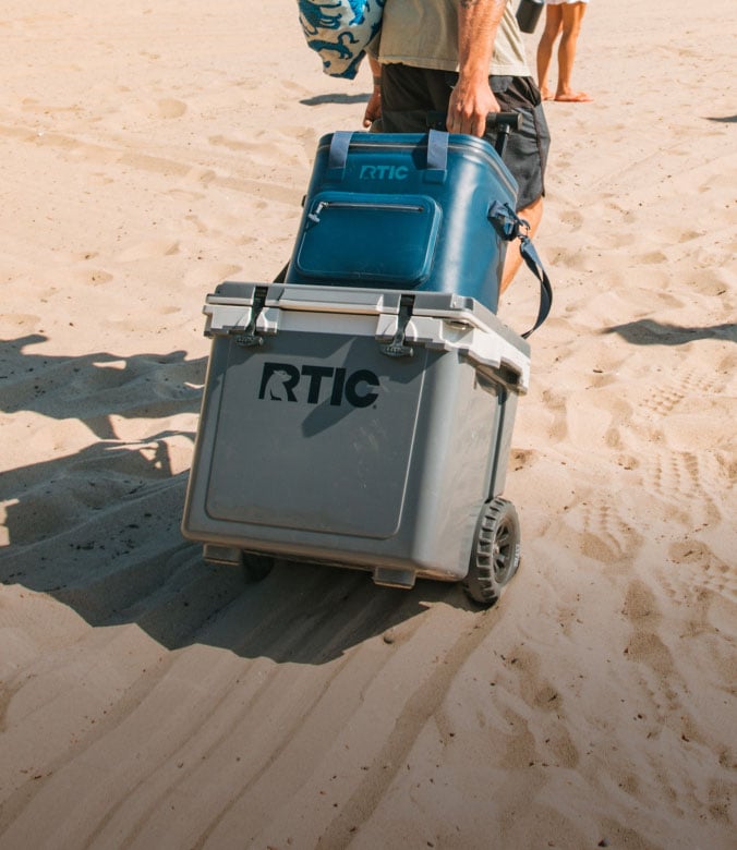 A person pulls a wheeled gray RTIC hard cooler stacked with a blue soft cooler bag across a sandy beach.