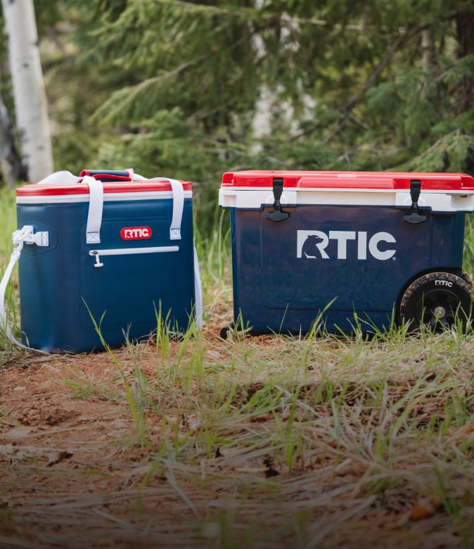 Red, white, and navy RTIC hard and soft cooler sitting on the forest floor. .