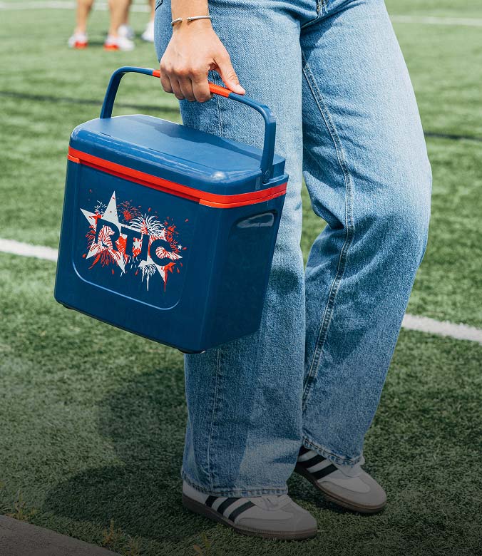 Person carrying a blue RTIC cooler with red accents across a sports field, wearing light blue jeans and casual sneakers.