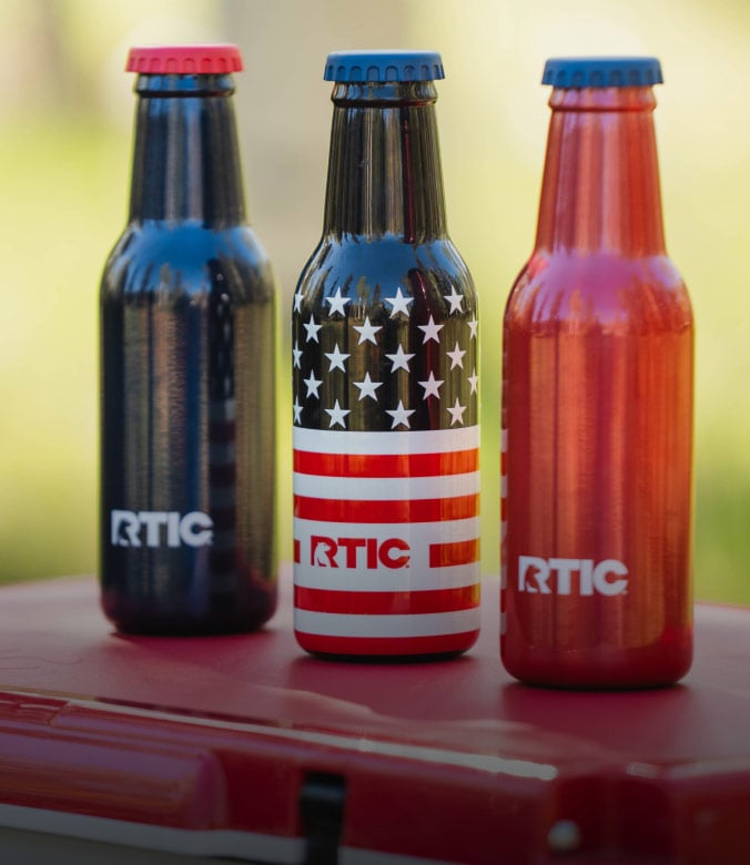 Three RTIC insulated bottles in patriotic red, white, and blue designs displayed outdoors on a red cooler lid.