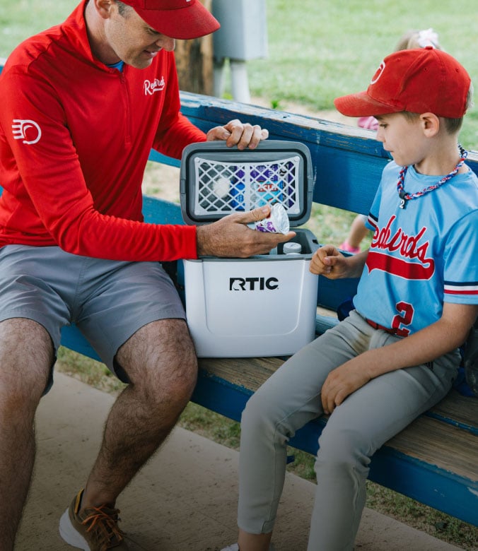Man handing a snack to a young baseball player from a white RTIC cooler during a game at the dugout.