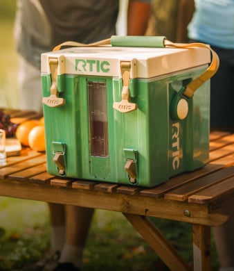 Green RTIC water cooler with latches on a wooden table outdoors.