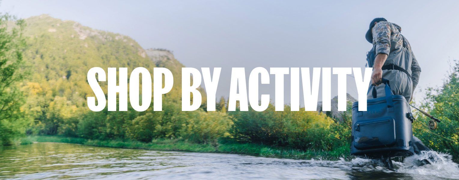 Person wading through a river carrying a blue RTIC Soft cooler in a mountain landscape, with “Shop by Activity” text overlaid on the image.