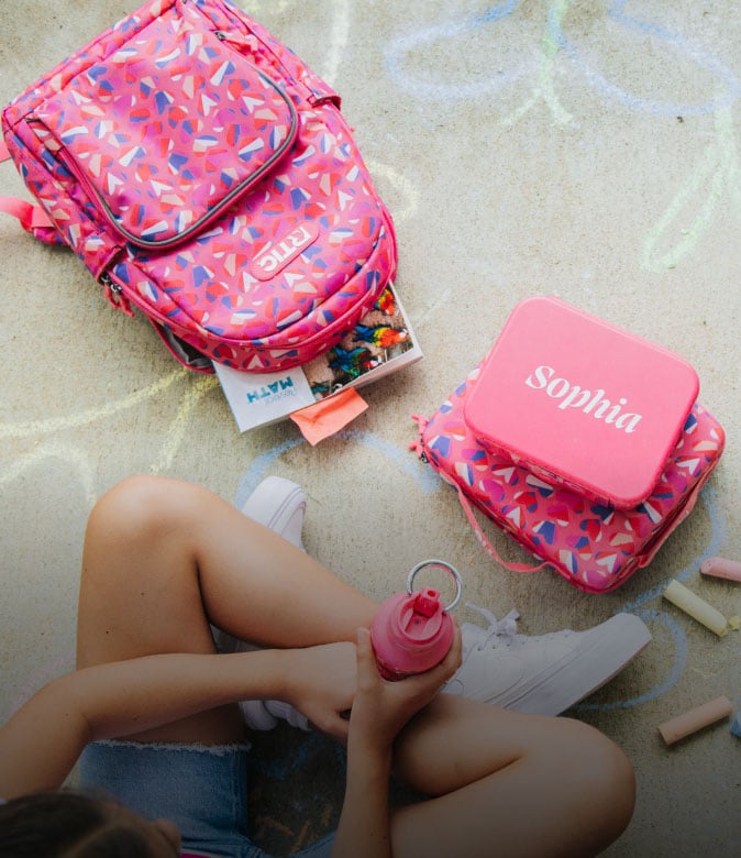 Pink patterned kids' backpack with matching lunch bag, water bottle, books, and chalk on a sidewalk.