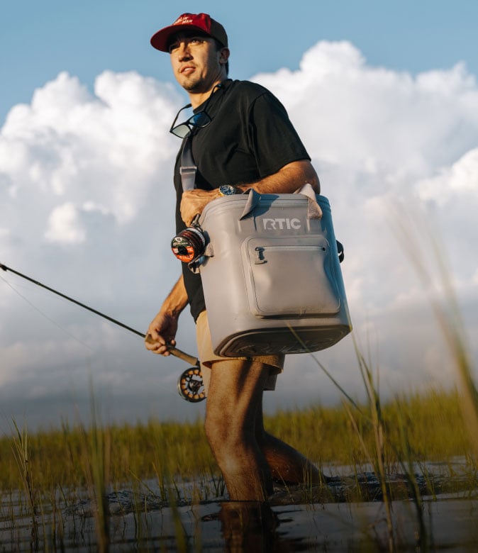 Man wading in a marshy wetland while carrying a gray RTIC soft cooler bag over his shoulder and holding a fishing rod