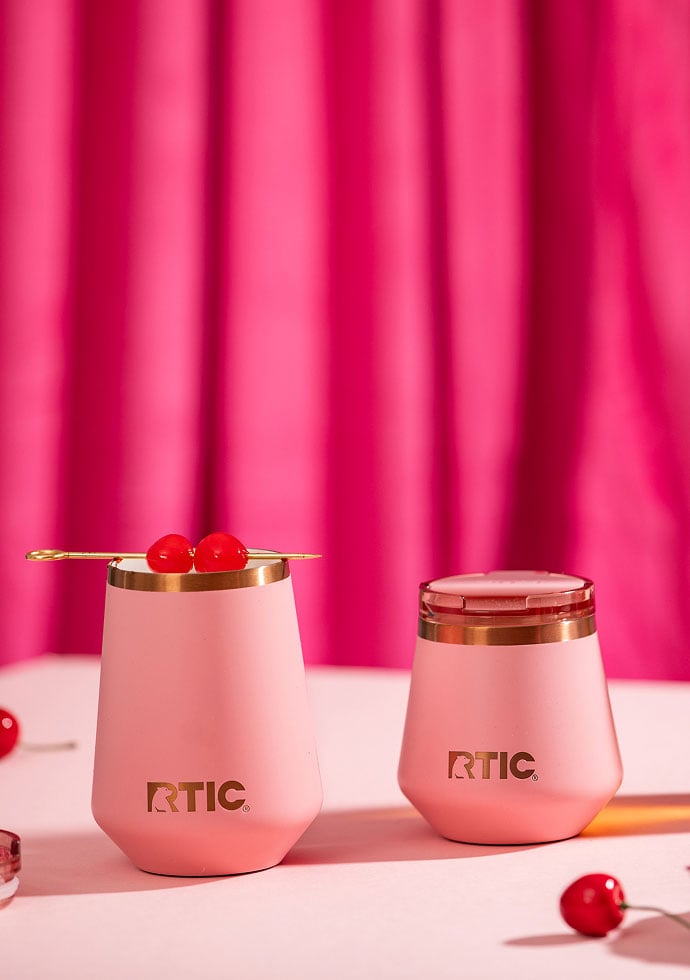 Two blush-pink RTIC insulated tumblers on a pink surface, one topped with a cocktail pick and cherries, against a bright pink backdrop.