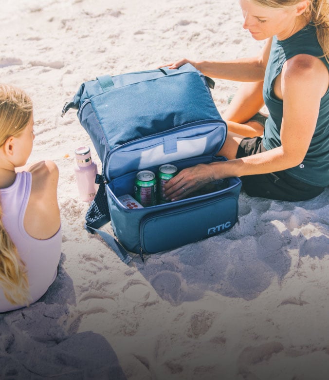 Woman opening a blue insulated cooler bag on the beach with canned drinks and snacks while a child sits nearby