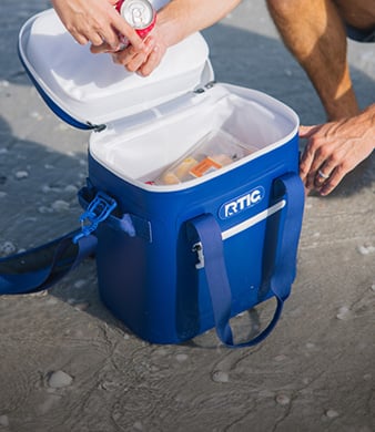 Blue RTIC soft cooler open on a sandy beach as two people place drinks inside for a day outdoors.