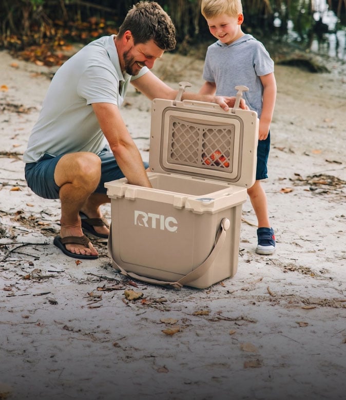 A man and a boy opening a tan RTIC hard cooler on the beach.