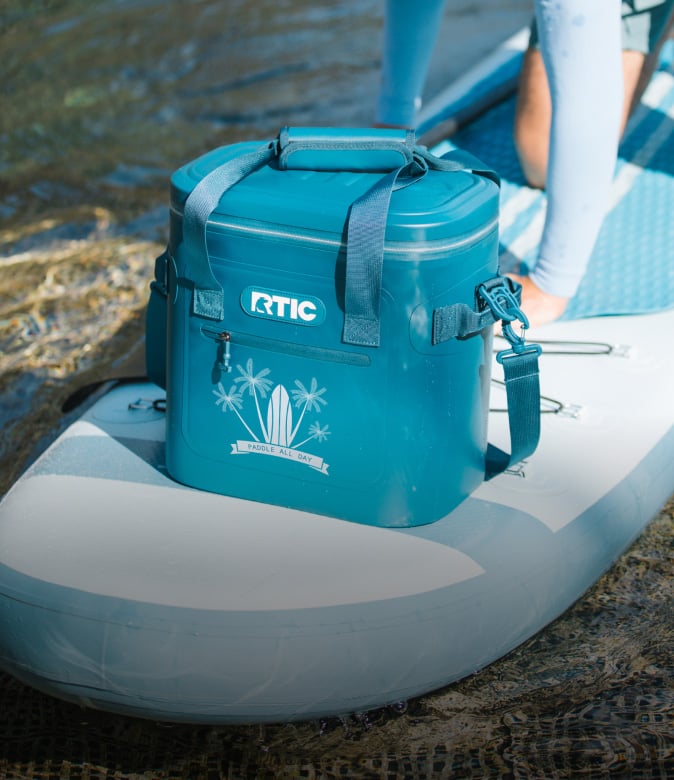 Blue RTIC soft cooler on top of an inflatable paddleboard on the water, featuring a “Paddle All Day” tropical graphic.