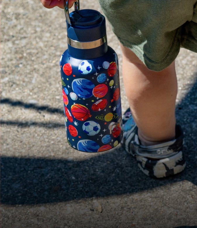 Child carrying a blue insulated water bottle designed with colorful planets and sports balls, walking outdoors on a sunny day.