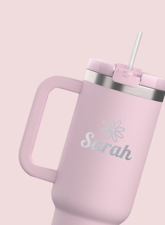 A light pink RTIC tumbler with a handle and a straw with the name Sarah and a flower engraved on it - on a light pink background.