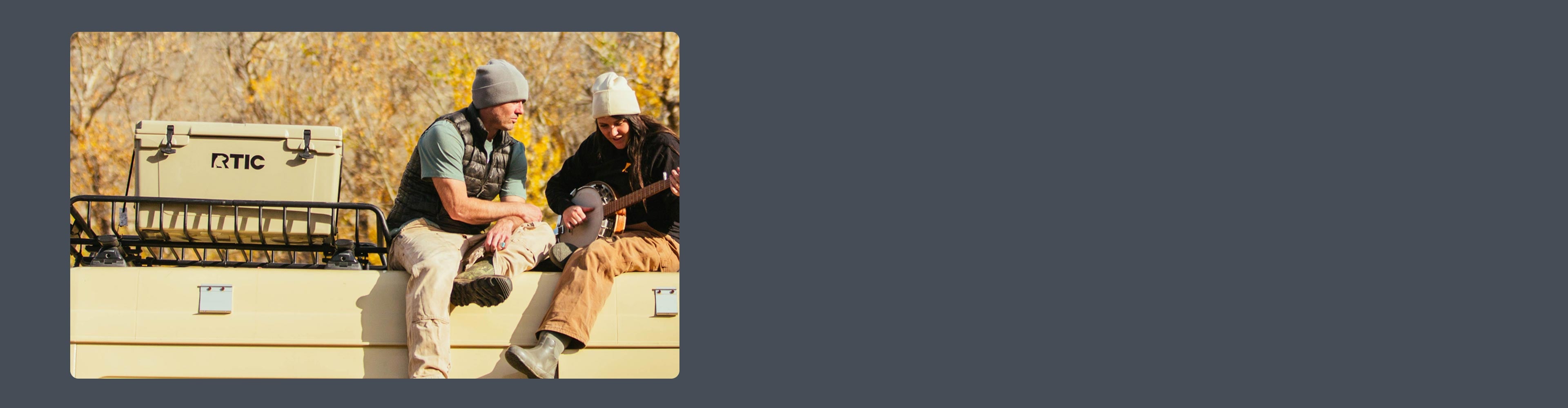 Two people sitting on the roof of a van, next to a RTIC Cooler; one is playing a banjo.