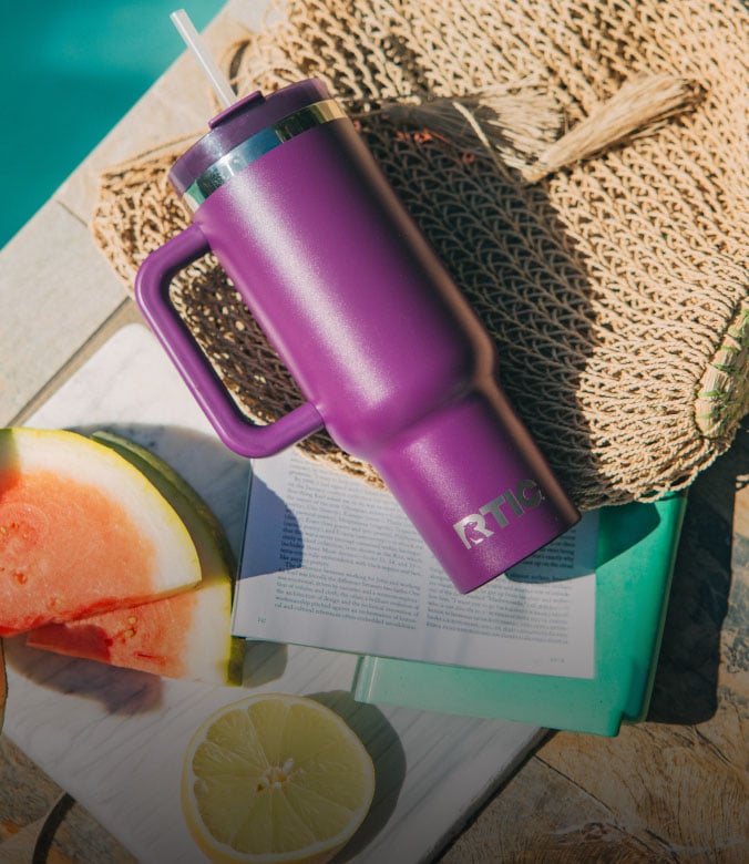 Purple RTIC insulated tumbler with handle and straw on a beach setup beside fruit, book, and woven bag.