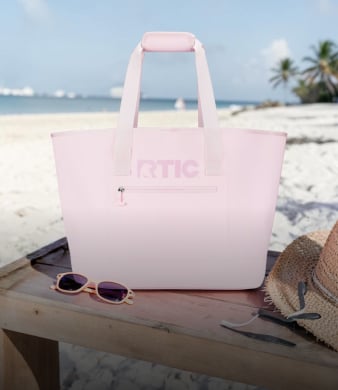Pink RTIC tote bag on a beach table with sunglasses and a hat nearby, ocean and palm trees in the background.