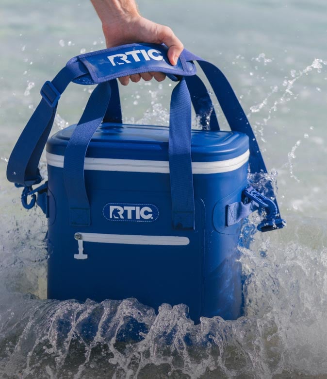 Hand lifting a blue RTIC soft cooler by its strap as waves splash around it in shallow water.
