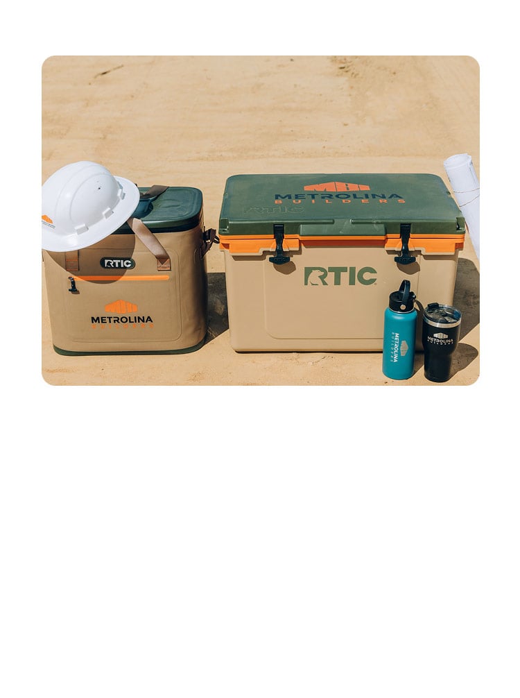 An RTIC Soft Cooler and RTIC Hard Cooler sitting on the dirt ground at a construction site, with a hard hat and water bottle next to them.