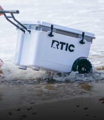 White RTIC wheeled cooler being pulled through shallow water at the beach, splashing as it rolls along the shoreline.
