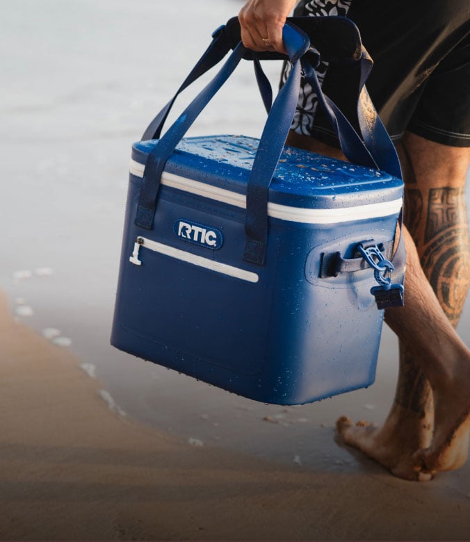 Close up of a person carrying a blue RTIC soft cooler on the beach.