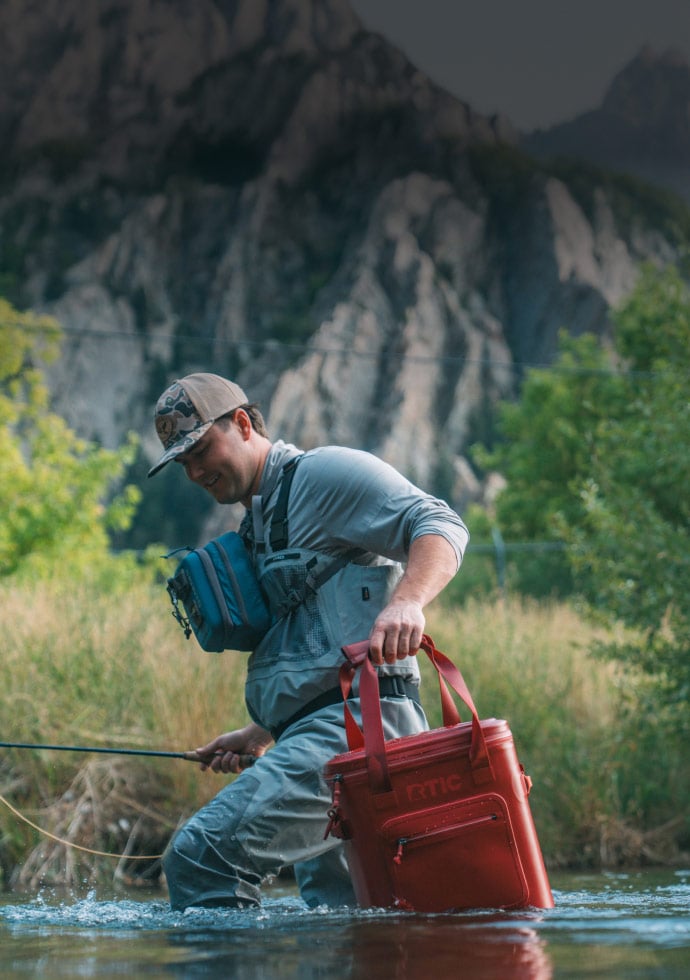 Angler wades through a river carrying a black cherry ultra-tough soft cooler pro and fishing rod, surrounded by trees and rocky canyon scenery.