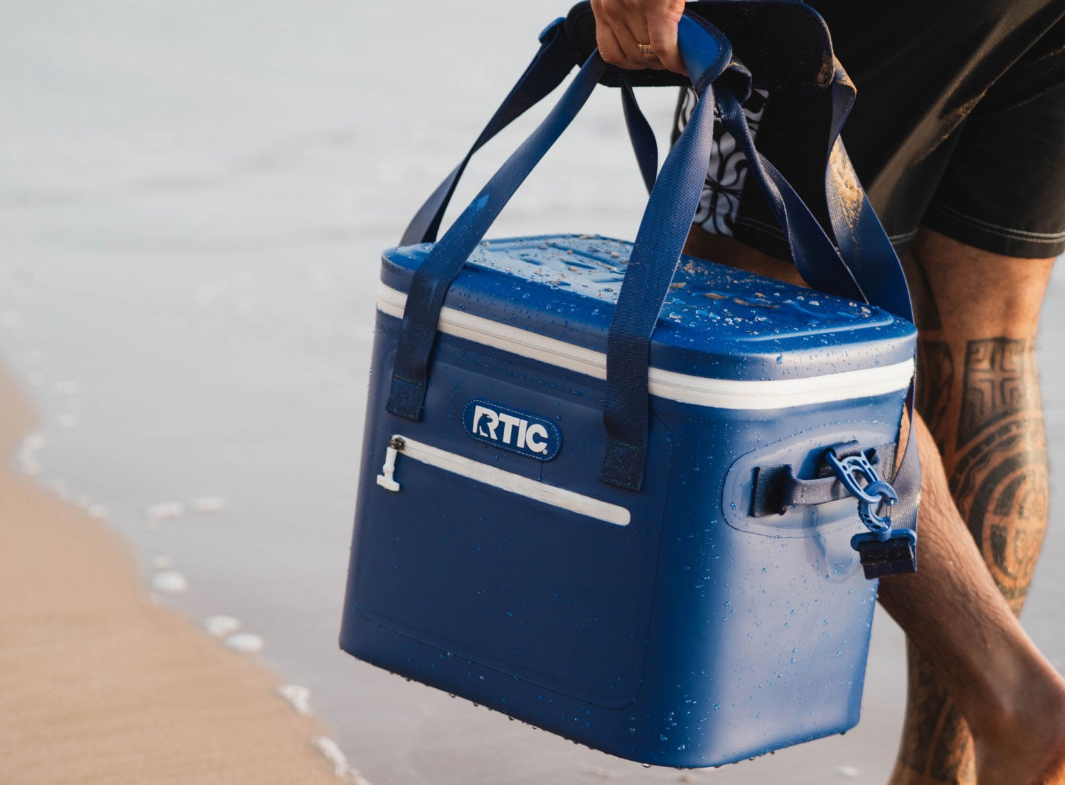 Close-up of a person walking on beach carrying a blue RTIC soft cooler.