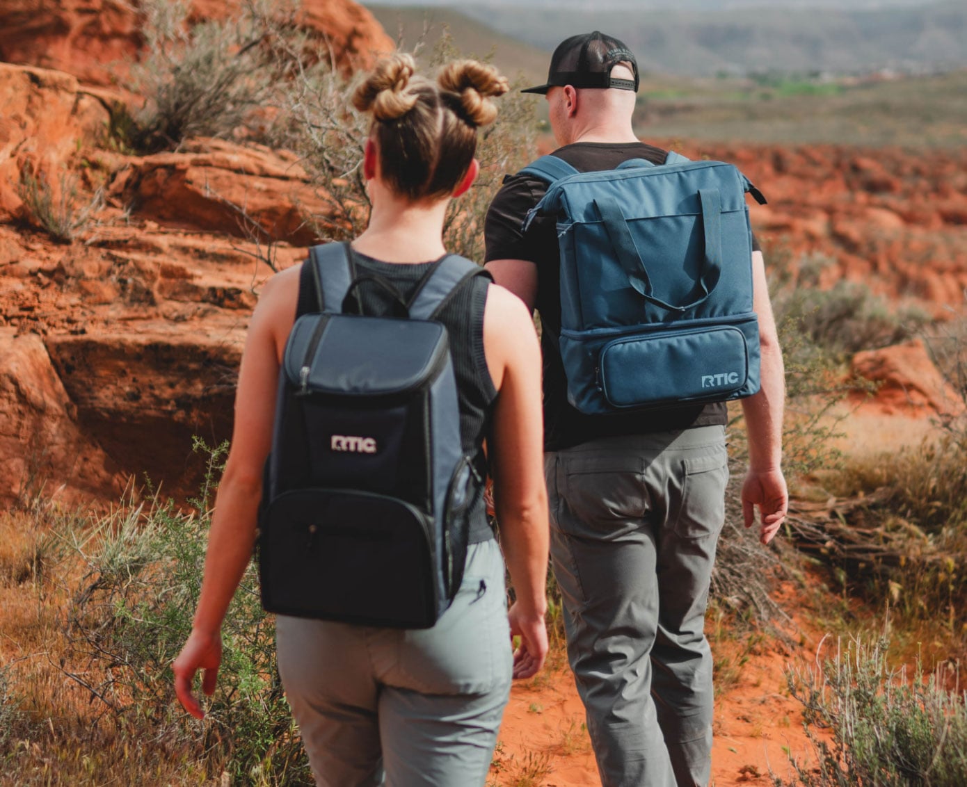 Two people hiking in a rocky desert landscape, wearing RTIC backpack coolers.