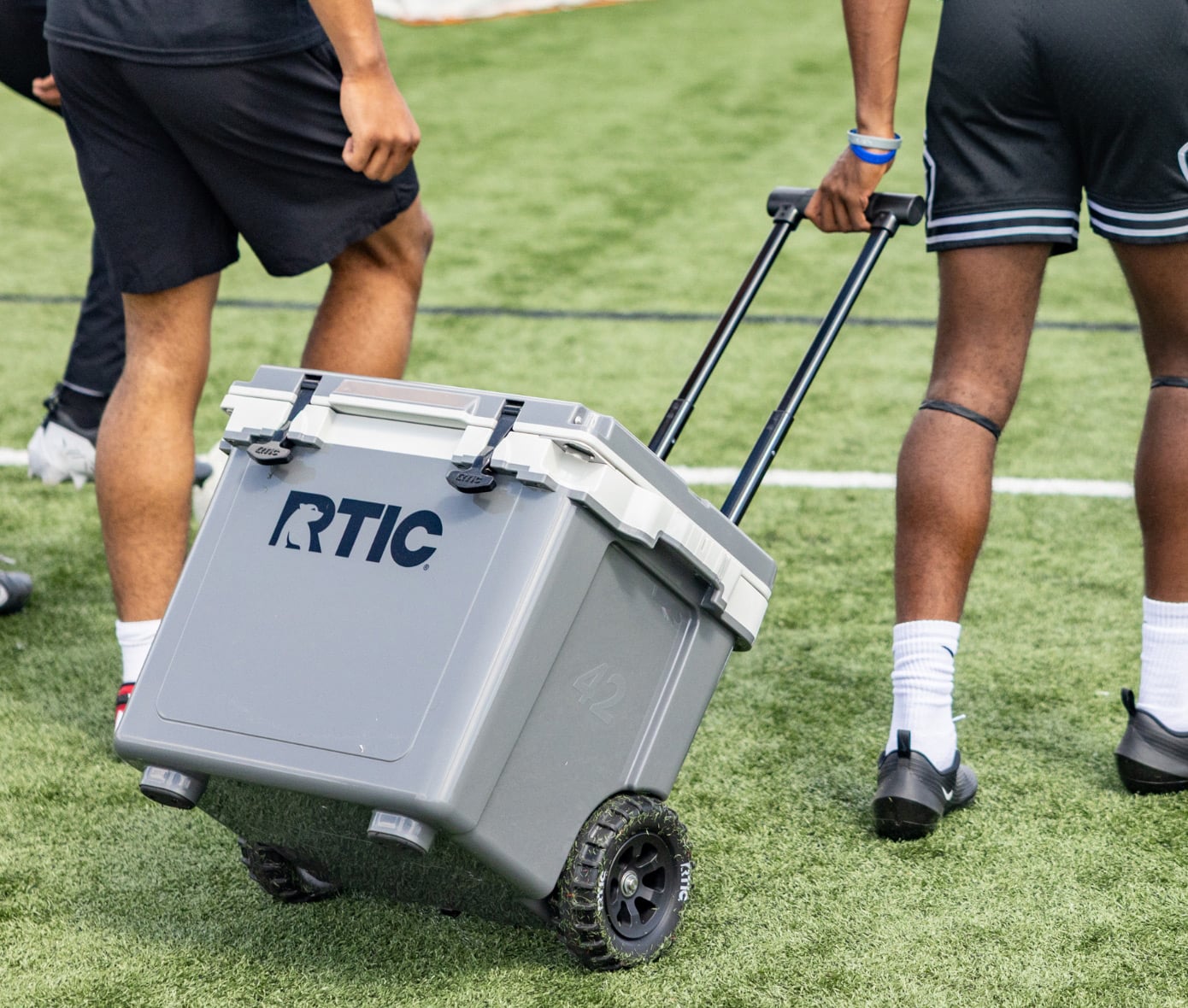 A person pulling a grey RTIC wheeled cooler on the grass soccer field.