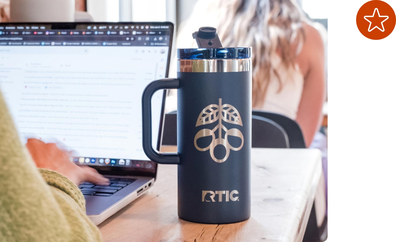 A customized navy blue RTIC mug on a table next to a person working on a laptop.