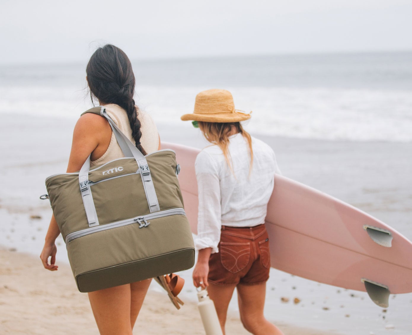 Two women walking on the beach, carrying an RTIC 2-in-1 Chillout Tote and a surfboard.